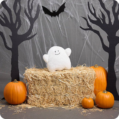 White ghost plush toy on a hay bale with pumpkins and Halloween decorations in the background.